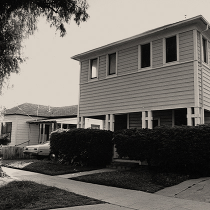The Harada House circa 1965, with dense hedges in front of the house, a tree by the sidewalk, and a car in the driveway.