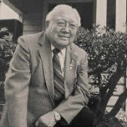 Harold Harada in a blue suit and red tie, kneeling in front of the Harada House, behind the National Historic Landmark plaque.