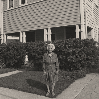 Sumi Harada in a dark dress, standing in the Harada House front yard, with dense hedges in front of the porch.