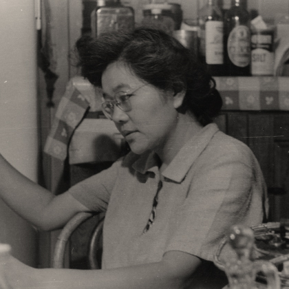 Sumi Harada seated at a table, reading the newspaper in the Harada House kitchen. Jars of bottles, condiments, and salt are visible on the cabinet behind.