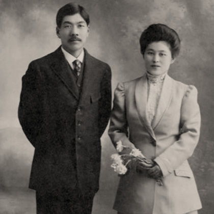 Jukichi Harada standing next to his wife Ken Harada. Jukichi wears a dark three piece suit, tie, and white shirt. Ken is holding flowers and is wearing gloves, a lace blouse, long Edwardian styled jacket, and long pleated skirt.