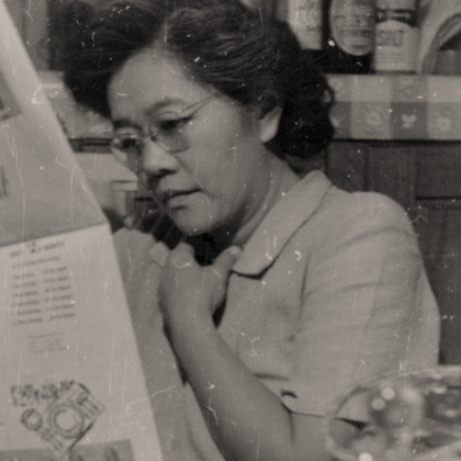 Sumi Harada seated at a table, reading the newspaper in the Harada House kitchen. Jars of bottles, condiments, and salt are visible on the cabinet behind.