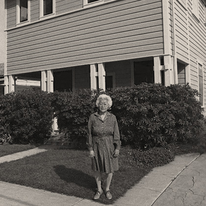 Sumi Harada in a dark dress, standing in the Harada House front yard, with dense hedges in front of the porch.