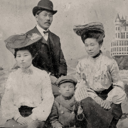 Ken, Jukichi, and Masa Atsu Harada with friend, photographed in front of a painted seaside resort backdrop. All are dressed in Edwardian attire. Masa Atsu is waving a US flag.