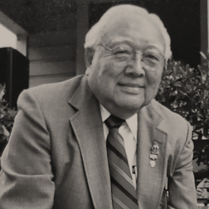 Harold Harada in a blue suit and red tie, kneeling in front of the Harada House, behind the National Historic Landmark plaque.