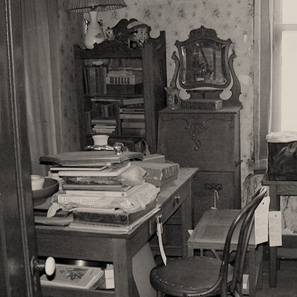 Antique furniture including a desk, secretary, and bentwood chair, each with a museum inventory tag. Books, papers, and boxes are stacked on the desk.