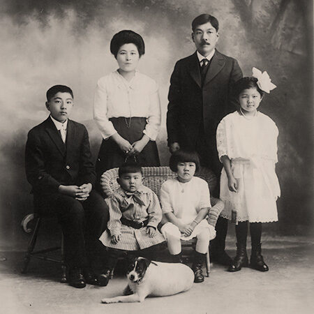 Formal portrait of the Harada family with family dog. Jukichi and Masa Atsu wear dark suits, Mine and Sumi wear white cotton dresses. Ken wears a white cotton blouse with dark skirt. Toddler Yoshizo wears a striped outfit.
